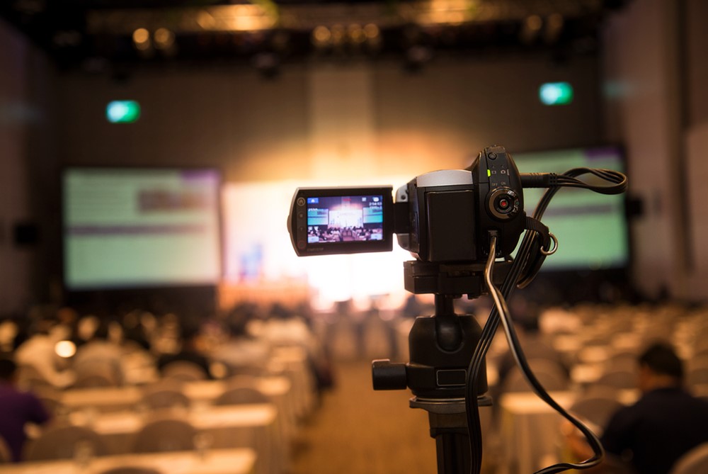 Closeup of a video camera set up in a big conference room.